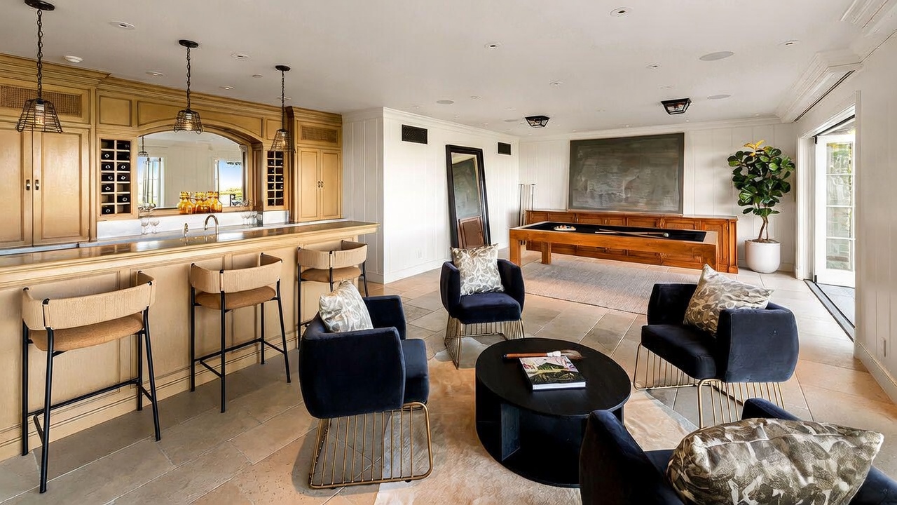 This spacious game room features a built-in wooden bar with pendant lighting, a pool table, and a comfortable seating area with dark blue armchairs and a circular coffee table. The room is finished with light-toned tile flooring and neutral walls, creating an inviting space for entertainment. Large glass doors provide natural light and a seamless transition to the outdoor area, while a large potted plant adds a touch of greenery to the corner.