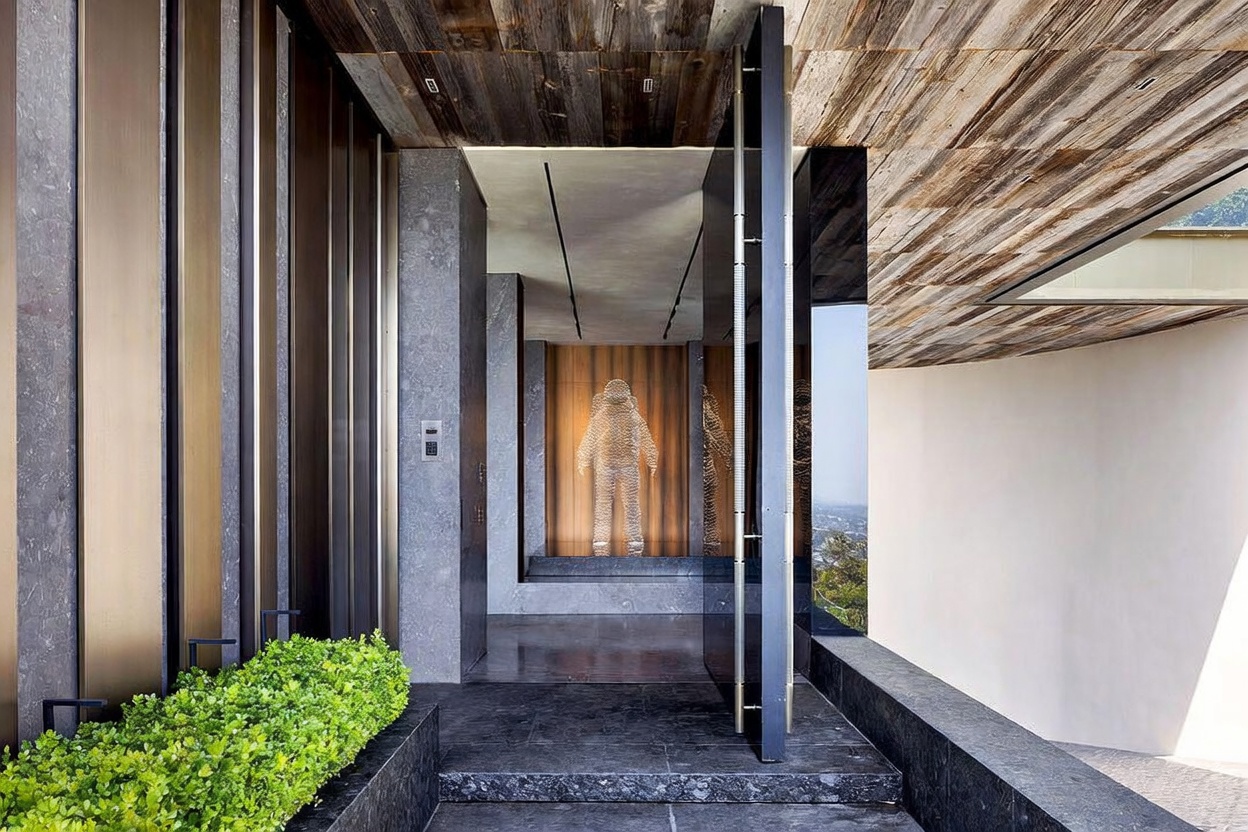 This striking modern entryway features a massive, floor-to-ceiling pivot door that opens into a sophisticated foyer adorned with a unique, illuminated wire-frame astronaut sculpture. The space is defined by a rich blend of textures, including reclaimed wood ceilings, dark stone flooring, and sleek metallic accents, creating a dramatic and luxurious first impression. The perspective looks directly through the threshold, highlighting the seamless transition between the exterior landscaping and the high-end interior design.