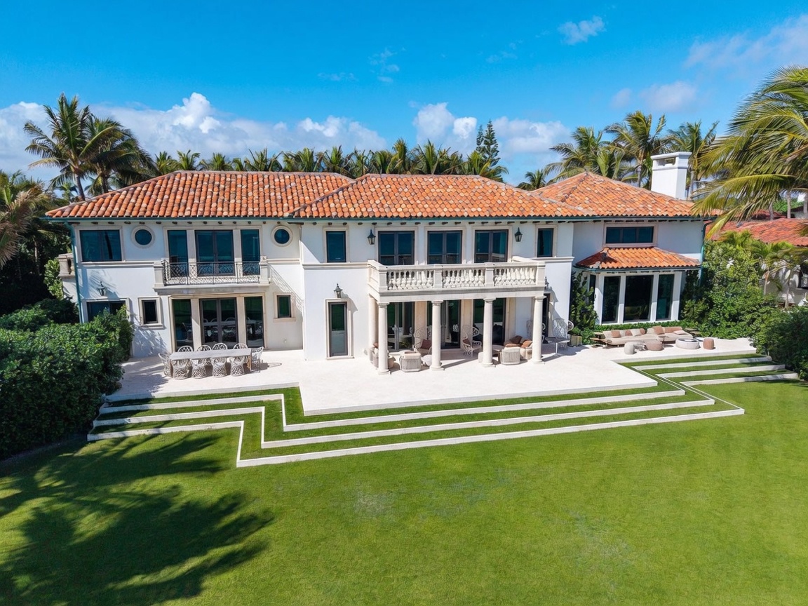 This expansive Mediterranean-style estate is captured from an elevated rear perspective, showcasing a symmetrical white stucco facade with a classic terracotta tile roof. The property features a sprawling stone patio with multiple seating areas, a grand colonnaded loggia, and a meticulously manicured lawn with tiered stone steps leading down from the terrace. The architecture is defined by its elegant proportions, numerous large windows, and a second-story balcony with decorative iron railings, all set against a bright blue sky.