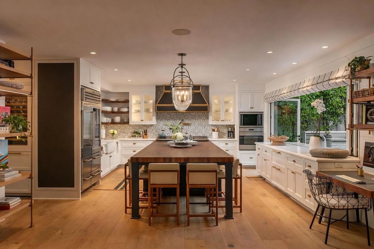 This sophisticated kitchen features a large central island with a dark wood top and integrated seating, serving as the focal point of the room. The space is defined by white cabinetry, a patterned tile backsplash, and a prominent range hood, all illuminated by recessed lighting and a decorative pendant fixture. Large windows with patterned shades open to an outdoor area, creating a bright and airy atmosphere with a seamless transition between indoor and outdoor living.