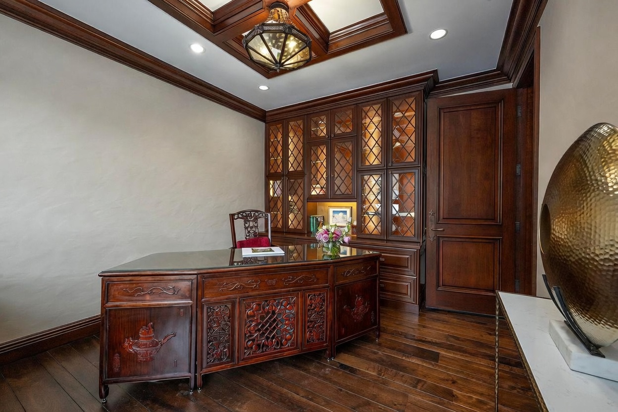 This sophisticated home office features rich, dark wood cabinetry with glass-paneled doors and intricate lattice detailing. A substantial, ornately carved wooden desk with a glass top serves as the centerpiece, complemented by a matching high-backed chair. The room is finished with dark hardwood flooring, a coffered ceiling with recessed lighting, and a decorative pendant fixture, creating an atmosphere of traditional elegance and professional focus.