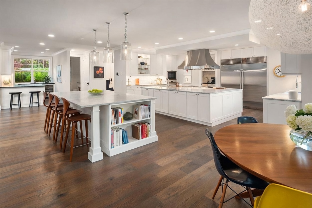 This expansive, open-concept kitchen features a large central island with a stone countertop and integrated bookshelf, complemented by a secondary island and white cabinetry. The space is illuminated by three glass pendant lights and recessed lighting, with warm-toned wood flooring grounding the bright, airy design. A dining area with a round table and modern chairs is visible in the foreground, creating a seamless transition between cooking and eating spaces.