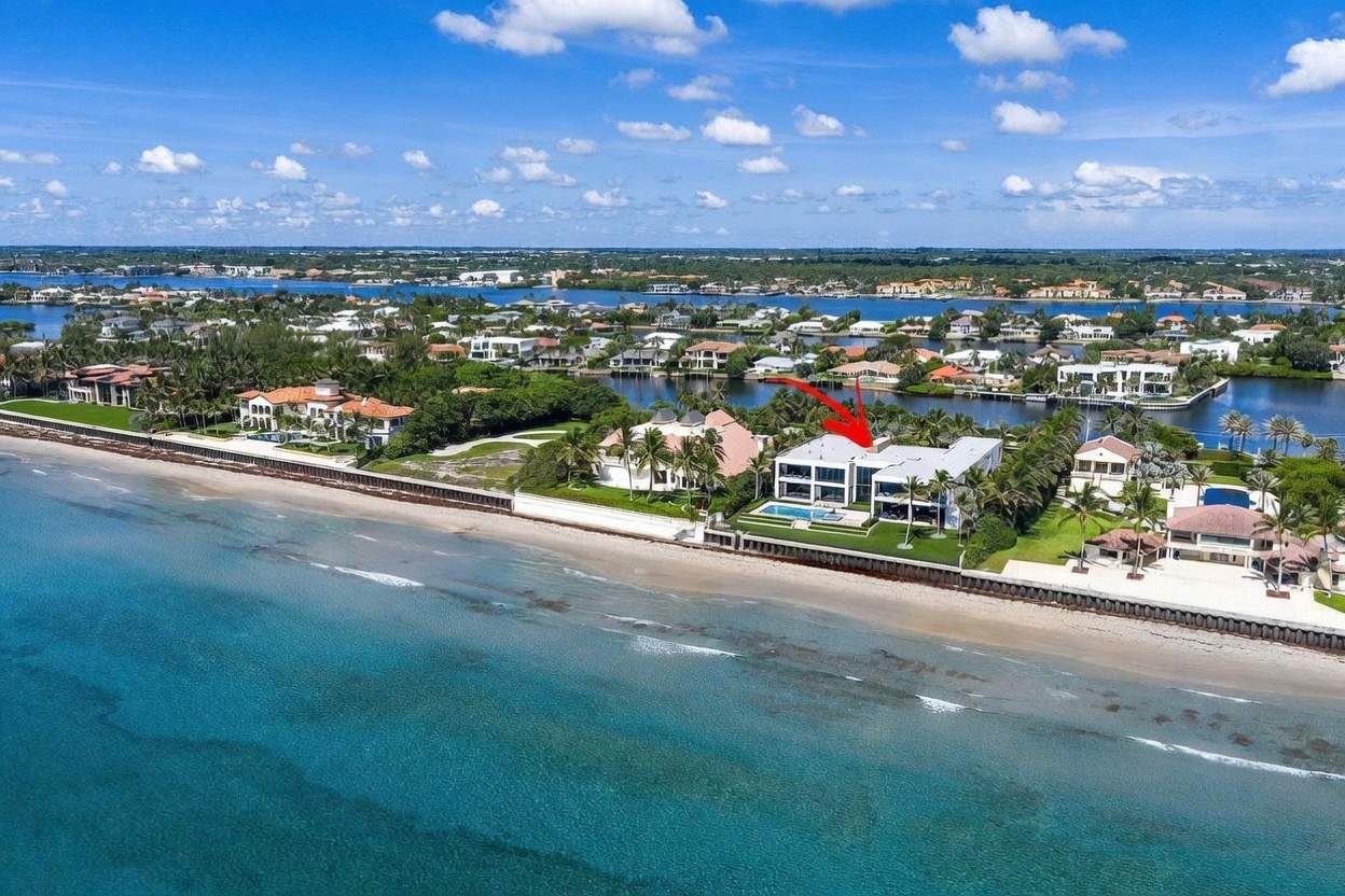 This high-angle aerial shot captures a luxurious beachfront estate situated along a pristine coastline. The modern residence features a flat roof, expansive glass walls, and a private swimming pool overlooking the ocean, with a red arrow highlighting the property. The surrounding landscape includes lush palm trees, manicured lawns, and a calm bay visible in the background, emphasizing the property's exclusive waterfront location.
