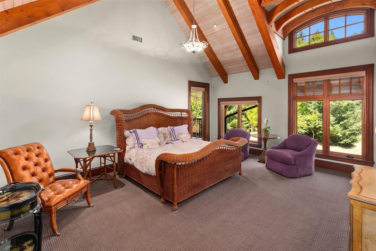 This spacious primary bedroom features a vaulted ceiling with exposed wooden beams and a central chandelier. The room is furnished with a large wicker-style bed frame, a tufted leather armchair, and two purple accent chairs arranged near large windows that provide ample natural light. The space is finished with neutral wall tones and wall-to-wall carpeting, creating a warm and inviting atmosphere.