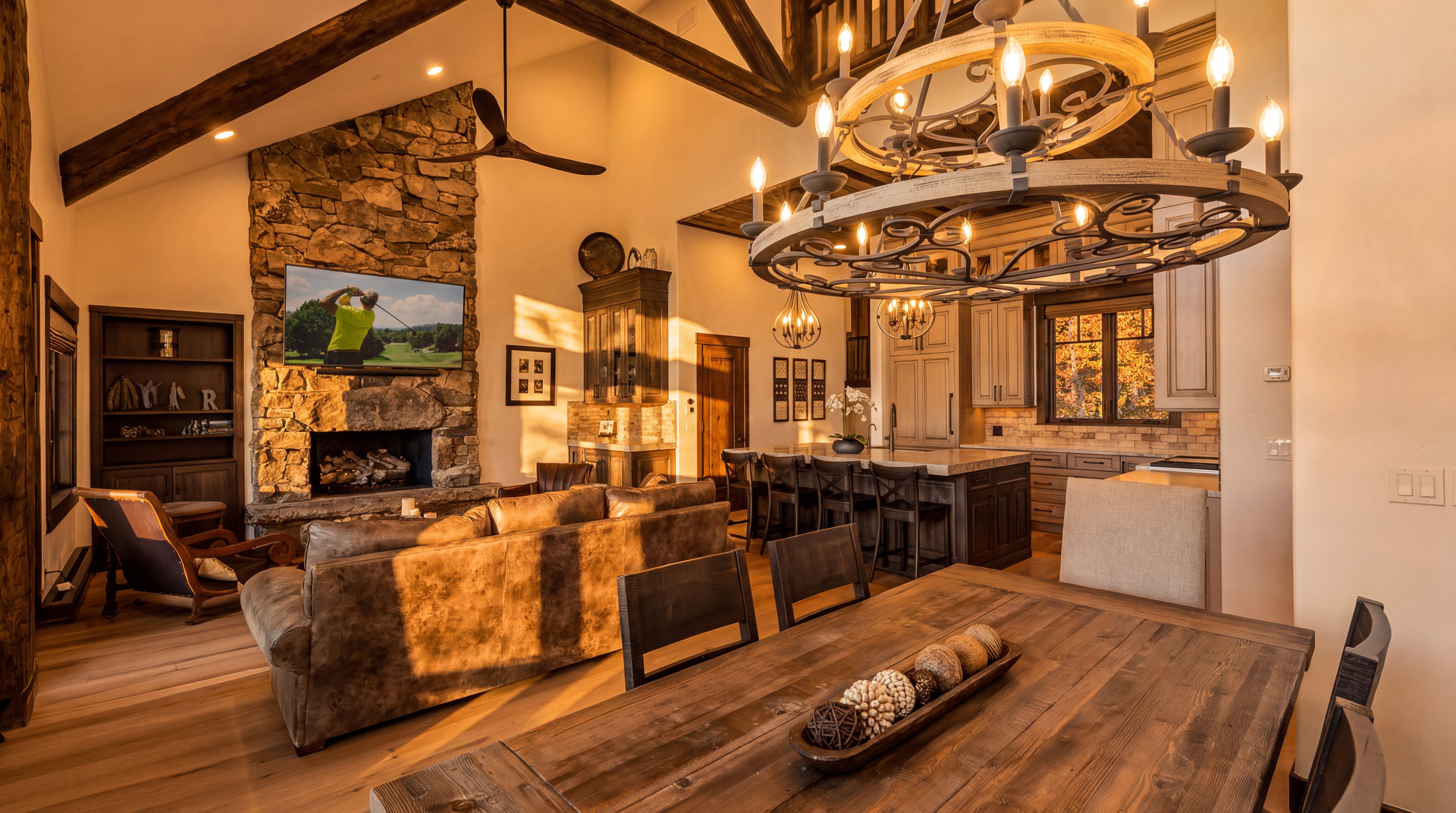 This spacious, open-concept floor plan seamlessly integrates a cozy living room, a dining area, and a gourmet kitchen, all featuring a warm, rustic aesthetic with exposed wooden beams and rich hardwood flooring. The living area is anchored by a stately stone fireplace with an integrated television, while the room is illuminated by a stunning, large circular wagon-wheel style chandelier. The rustic, mountain-inspired decor is accentuated by heavy leather furnishings and natural tones, creating a welcoming and sophisticated home environment.