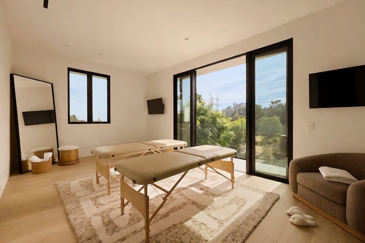 This interior space is designed as a professional massage or wellness room, featuring two portable massage tables centered on a textured area rug. The room is minimalist and bright, with light wood flooring, white walls, and a large glass door that opens to an outdoor balcony with a view of trees. A tall floor mirror, a small stool, and a comfortable armchair complete the serene, spa-like atmosphere.