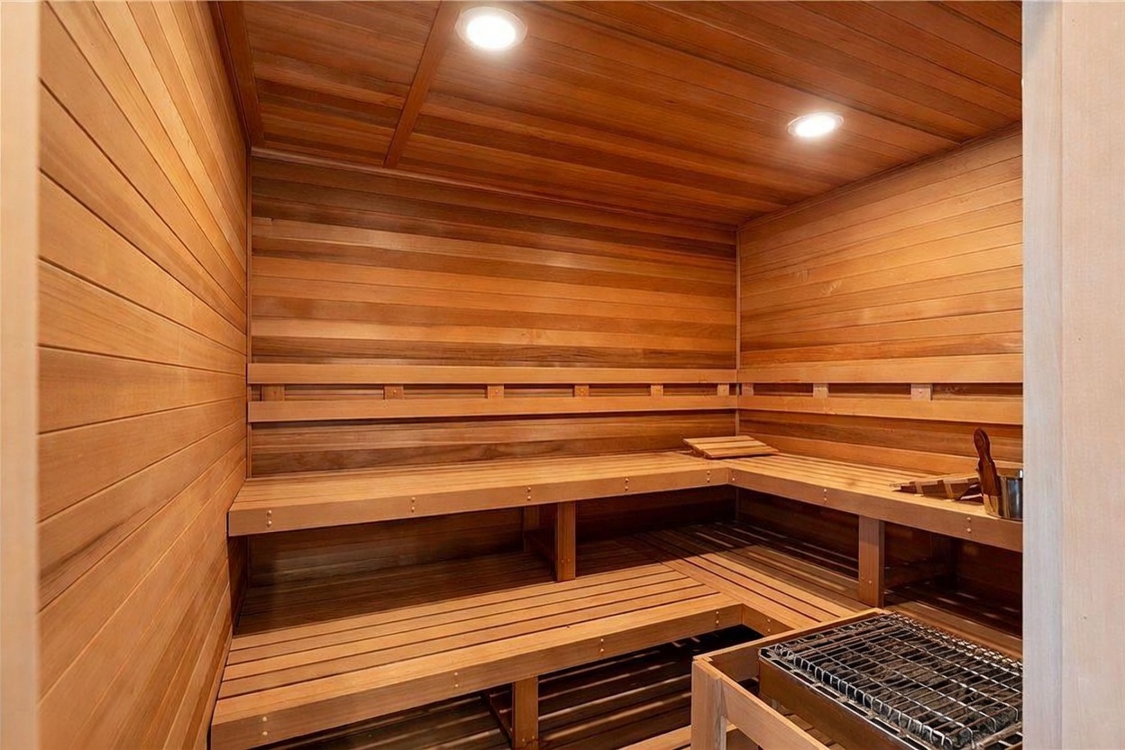 This interior view showcases a custom-built wooden sauna featuring tiered bench seating and warm, recessed lighting. The space is lined with horizontal wood paneling, creating a cohesive and relaxing atmosphere. A metal heater unit with stones is positioned in the foreground, completing the authentic sauna experience.