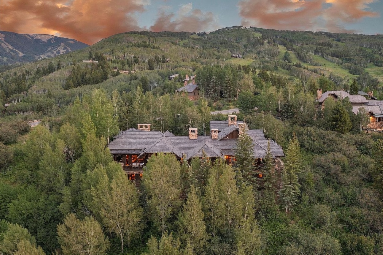 This aerial perspective captures a sprawling mountain estate nestled within a dense forest of aspen and pine trees. The home features a complex, multi-gabled roofline with stone and wood siding, blending seamlessly into the rugged, elevated landscape. The warm glow from the interior lights contrasts beautifully against the dramatic, sunset-lit sky and the surrounding natural terrain.