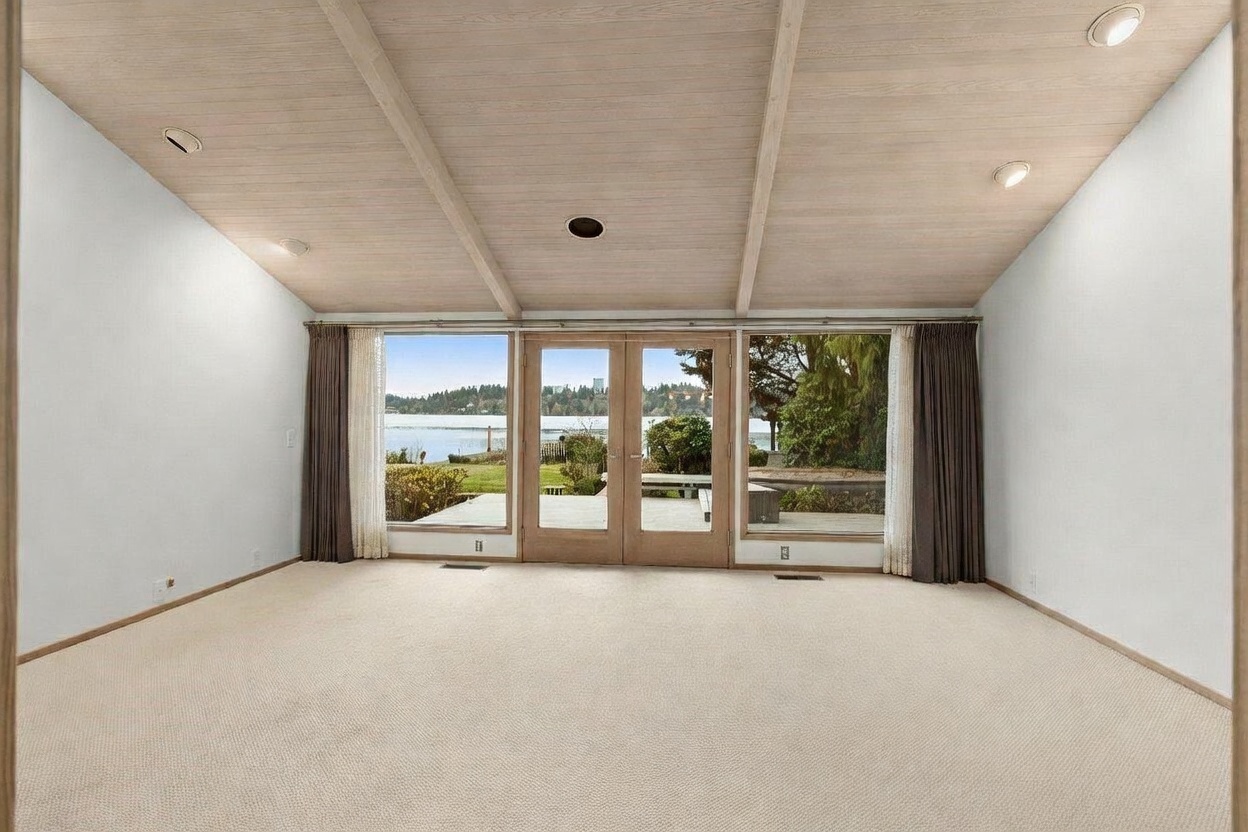 This spacious living room features a vaulted, light wood-paneled ceiling and neutral wall-to-wall carpeting. A central set of French doors flanked by large windows provides a direct view of the lake and outdoor landscape. The room is framed by dark curtains on either side, creating a balanced and serene atmosphere with a focus on the natural scenery.