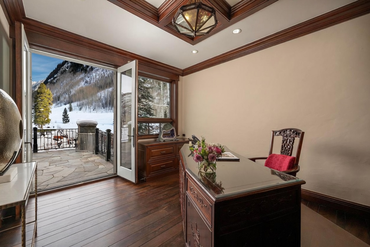 This elegant home office features rich wood paneling, a coffered ceiling with a decorative lantern-style chandelier, and dark hardwood flooring. A large glass-topped desk sits in the foreground, while an open door leads directly to a stone-paved balcony overlooking a snowy mountain landscape. The room exudes a sophisticated, professional atmosphere with high-end finishes and a seamless connection to the outdoors.