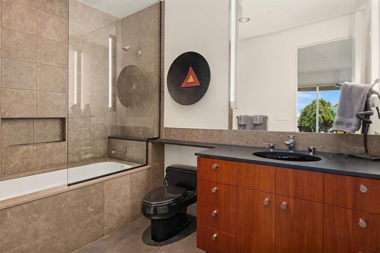 This modern bathroom features a sleek, dark-toned vanity with a black sink and wood-finish cabinetry, complemented by a black toilet. A glass-enclosed shower and tub combination is clad in neutral-toned stone tiles, creating a cohesive and contemporary aesthetic. A large mirror reflects the space, including a view of an outdoor area, while a unique circular wall art piece adds a bold decorative touch.