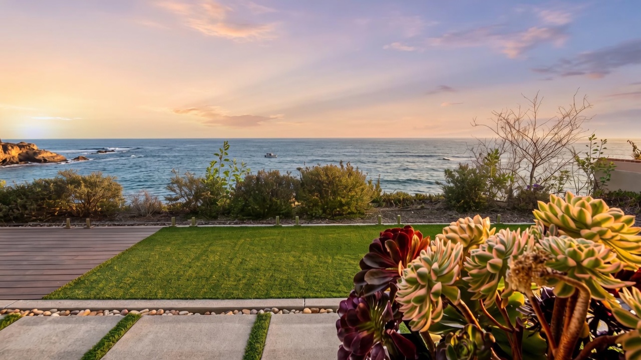This image captures a serene outdoor living space featuring a wooden deck and a patch of manicured artificial turf that overlooks the ocean. A glass railing provides an unobstructed view of the coastline, while a large potted succulent arrangement in the foreground adds a touch of natural texture. The perspective is from a slightly elevated position, emphasizing the seamless transition between the private patio and the expansive coastal horizon.