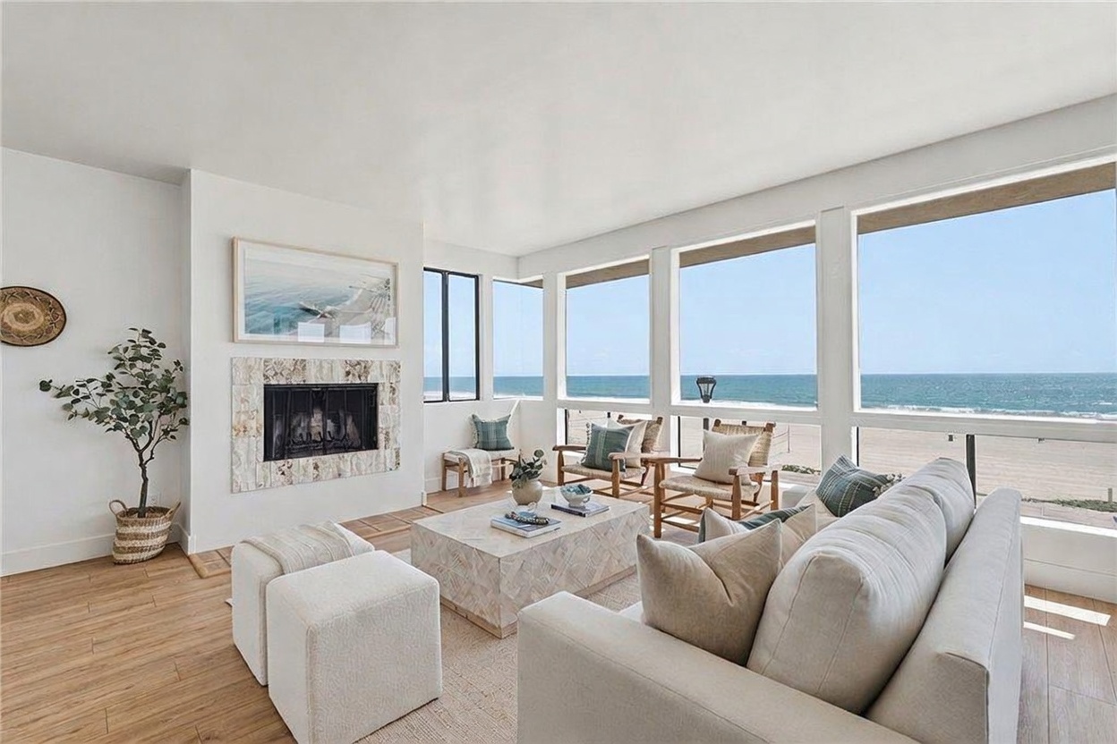 This bright and airy living room features a neutral color palette and light wood flooring, creating a serene coastal atmosphere. A large, light-toned sofa and a central stone-topped coffee table face a fireplace with a textured stone surround, while expansive windows provide unobstructed, panoramic views of the ocean. The space is thoughtfully furnished with woven chairs and decorative accents, emphasizing a relaxed, upscale beachside lifestyle.