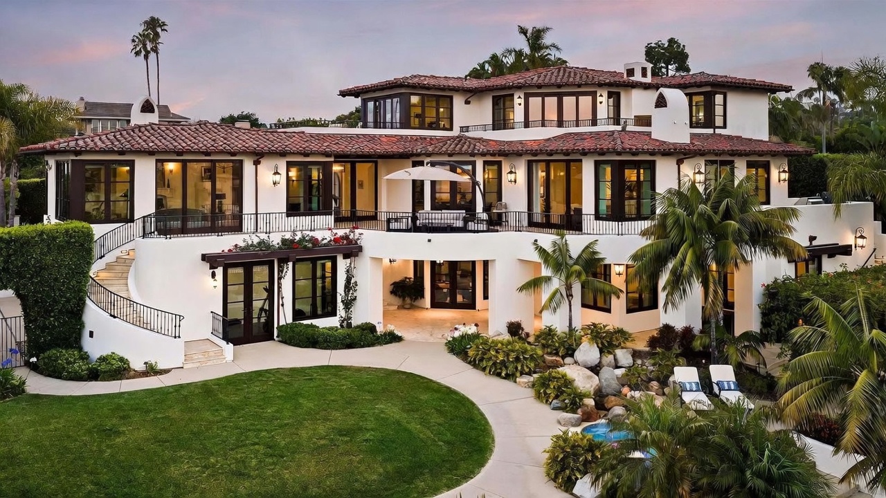 This expansive Mediterranean-style estate is captured from a rear perspective, showcasing a multi-level design with a classic red-tiled roof and white stucco exterior. The property features a large upper-level balcony with wrought-iron railings, a curved staircase leading to the lawn, and a covered lower-level patio area that opens onto a landscaped backyard with a rock-accented water feature. The scene is set during the golden hour, highlighting the warm lighting throughout the home and the lush, manicured greenery.