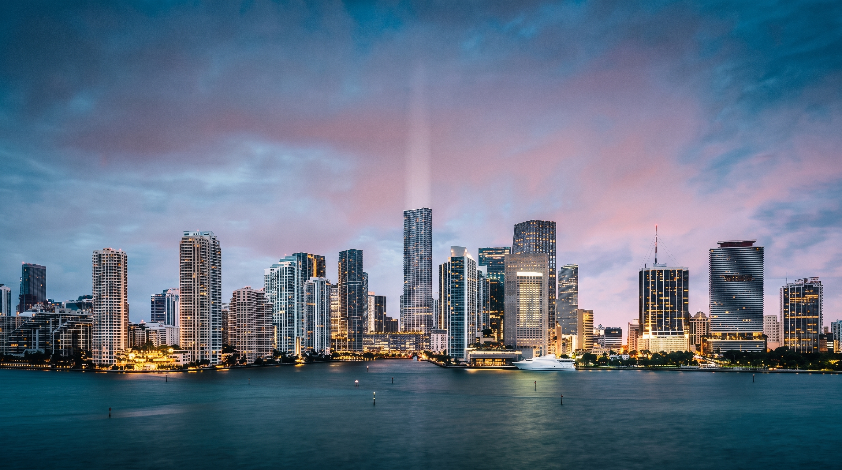 This wide-angle, cinematic shot captures the expansive Miami skyline at dusk, viewed from across the water. The dense cluster of high-rise residential and commercial towers creates a dramatic silhouette against a soft, pastel-hued sky. The calm water in the foreground reflects the fading light, emphasizing the prime waterfront location and the vibrant urban lifestyle of the area.