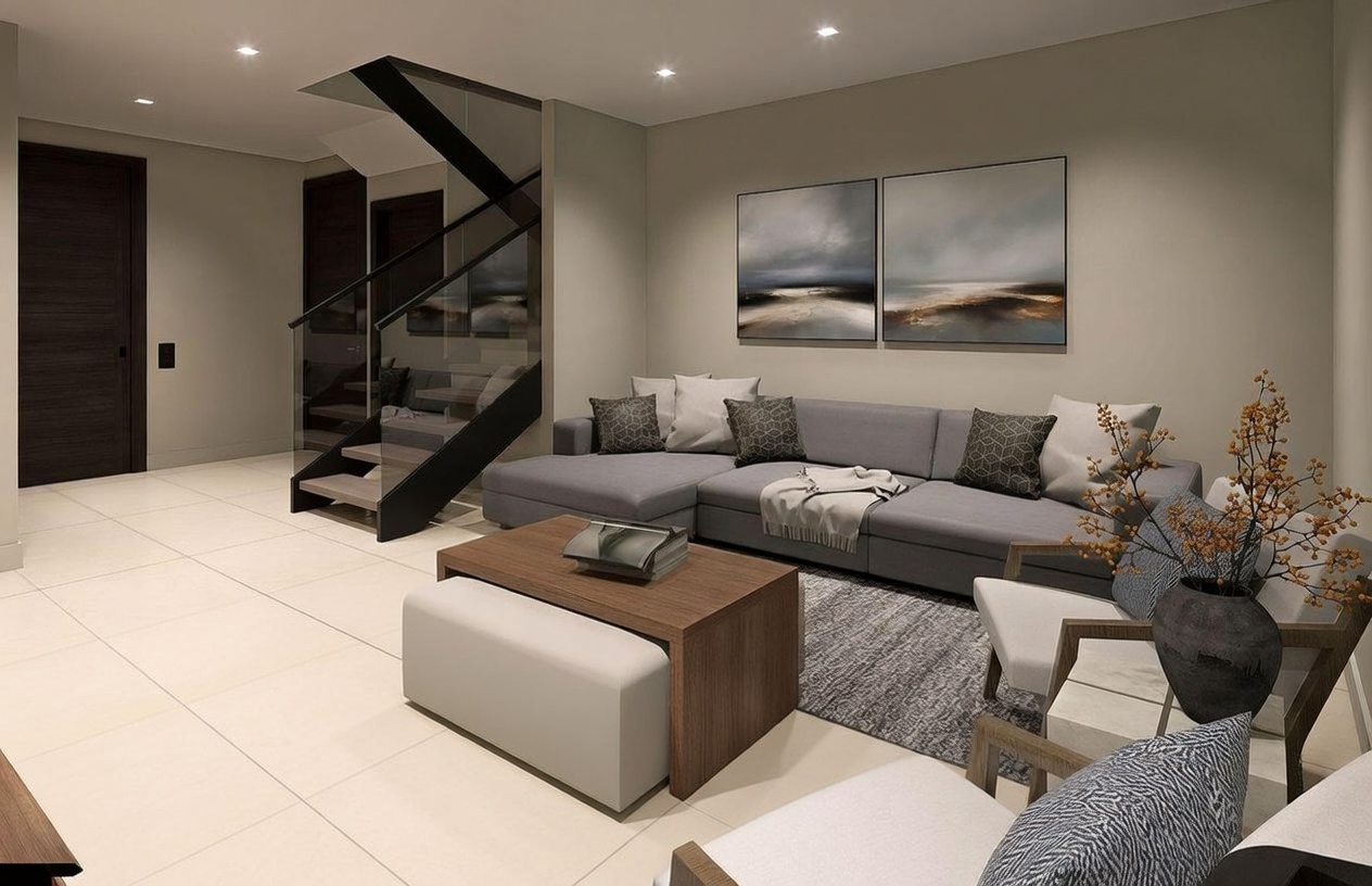 This contemporary living room features a large grey sectional sofa accented with patterned throw pillows and a neutral-toned area rug. A central wooden coffee table with a matching ottoman provides a functional focal point, while two modern armchairs offer additional seating. The space is illuminated by recessed ceiling lights and includes a striking staircase with glass railings, creating an open and sophisticated atmosphere.
