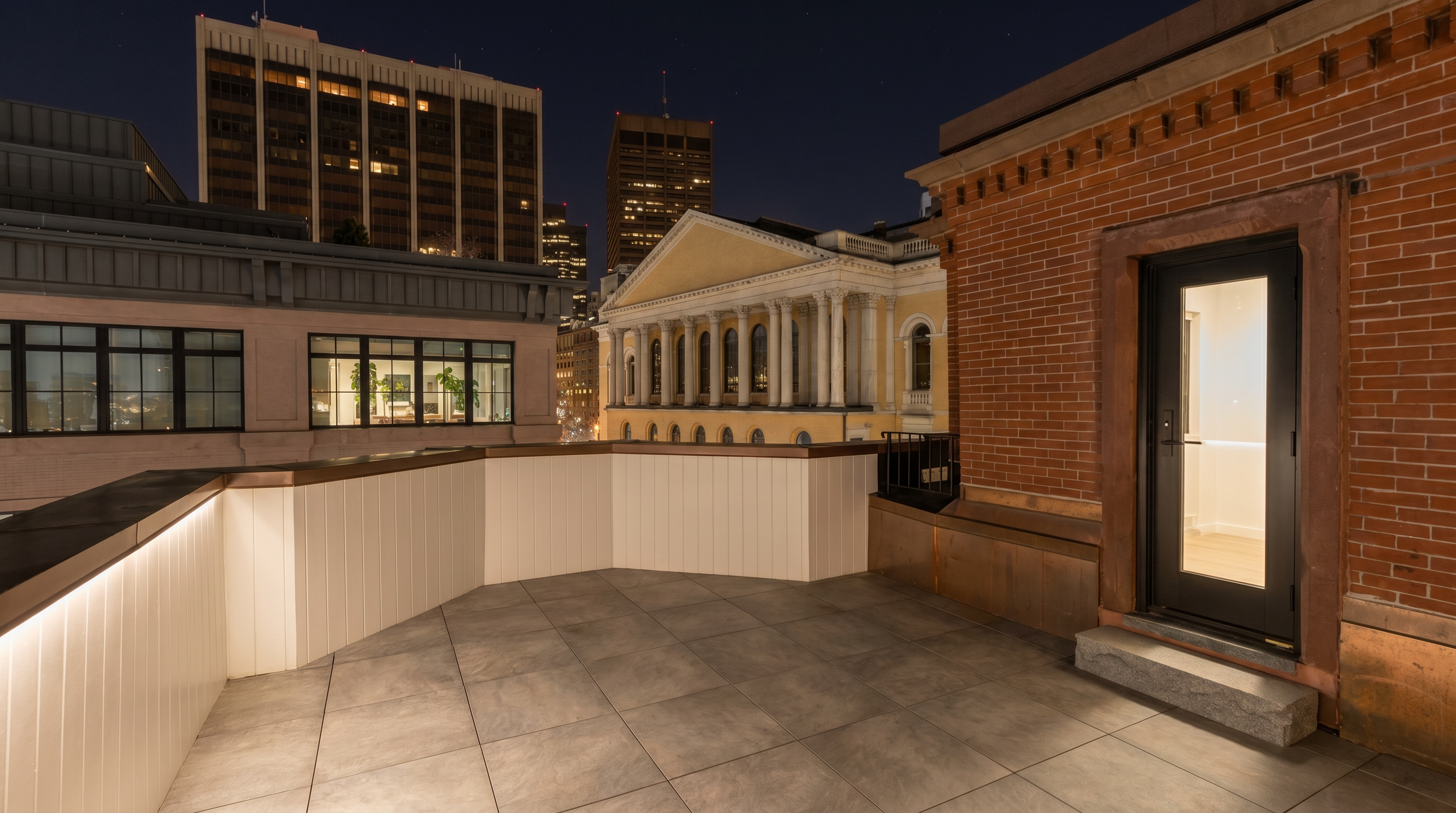 This rooftop terrace features modern, large-format gray tiles and a clean white railing, providing a private outdoor space. The area is accessed through a sleek black-framed glass door set into a classic red brick wall. The perspective offers an elevated view of the surrounding urban architecture, including a historic building with classical columns and modern high-rise structures in the distance.