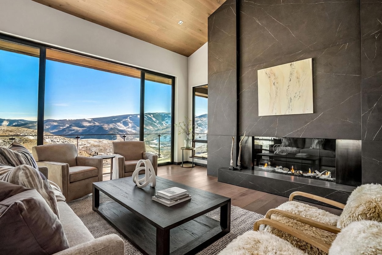 This is a luxurious living room featuring a modern fireplace with a dark marble surround and a contemporary art piece above. Large windows offer stunning mountain views, and the room is furnished with comfortable seating and a dark wood coffee table. The overall aesthetic is sophisticated and inviting, perfect for relaxation and enjoying the scenery.