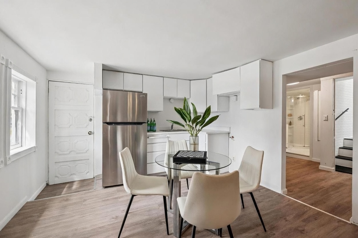 This bright, open-concept kitchen and dining area features white cabinetry, stainless steel appliances, and light wood-look flooring. A glass-topped dining table with four modern chairs sits in the center of the room, accented by a decorative plant. The space flows seamlessly into an adjacent hallway, providing a clean and functional layout.