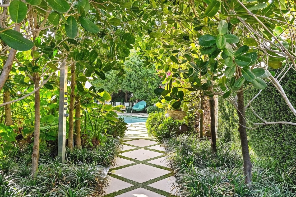 A lush, tropical garden path leads toward a sparkling swimming pool, framed by mature trees and dense greenery. The walkway features a geometric pattern of stone pavers set within artificial turf, creating a modern and manicured aesthetic. A sleek, metallic outdoor shower stands to the left, while a vibrant blue lounge chair is visible in the distance, inviting relaxation in this private outdoor oasis.