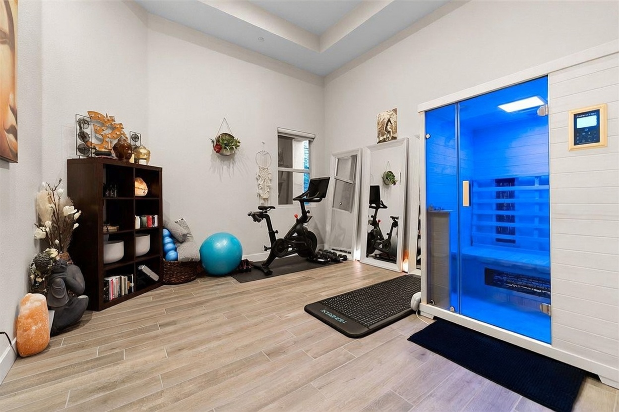 This home gym features light-toned wood-look tile flooring and a dedicated wellness area equipped with a stationary exercise bike and a blue-lit infrared sauna. A large floor mirror leans against the wall, while a shelving unit displays decorative items and sound bowls, creating a serene and functional workout space. The room is brightly lit with recessed lighting, offering a clean and modern aesthetic suitable for fitness and relaxation.