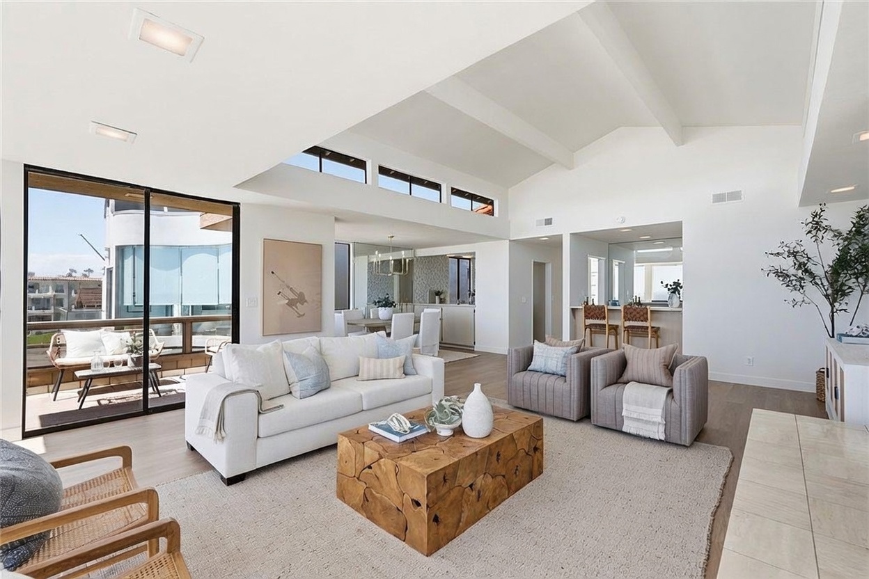 This bright and airy living room features a vaulted ceiling with exposed beams and a series of clerestory windows that flood the space with natural light. The room is furnished with a white sofa, two gray armchairs, and a distinctive natural wood coffee table, all arranged on a large textured area rug. A sliding glass door provides a seamless transition to an outdoor deck, while the open floor plan connects the living area to a dining space and a kitchen bar in the background.