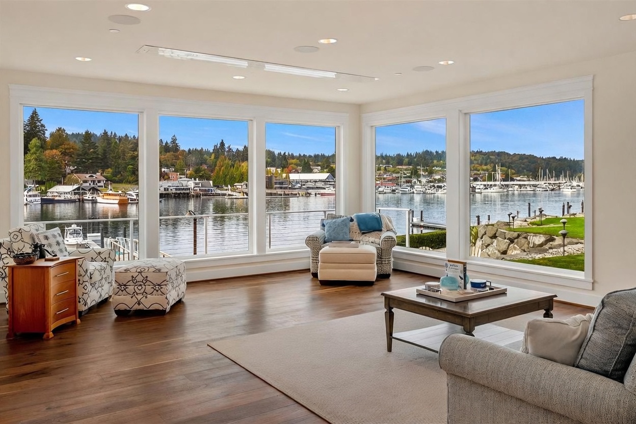 This bright and airy living room features expansive windows that provide a panoramic view of a tranquil harbor filled with boats. The space is furnished with comfortable patterned armchairs, a plush ottoman, and a central coffee table resting on a neutral area rug, all set against rich wood flooring. The room's open layout and abundant natural light create a serene and inviting atmosphere perfect for waterfront living.