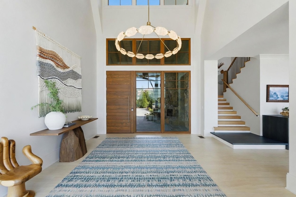 This grand entryway features a soaring vaulted ceiling and a striking circular chandelier that serves as a focal point. A large wooden front door is flanked by glass panels, allowing natural light to pour into the space, while a textured wall hanging and a console table add a touch of organic warmth. To the right, a staircase with a wooden railing leads to the upper level, set against a backdrop of clean white walls and light-toned flooring.