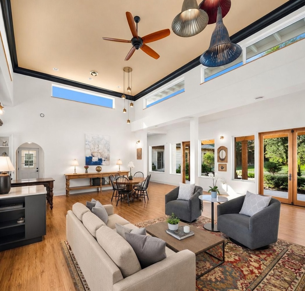 This spacious, open-concept living and dining area features high ceilings with a dark trim accent and a central wooden ceiling fan. The room is bathed in natural light from multiple windows, including high-placed clerestory windows, and includes a comfortable seating area with a patterned rug and a separate dining space with a long wooden console table. The neutral color palette and warm wood flooring create an inviting, airy atmosphere suitable for both relaxation and entertaining.