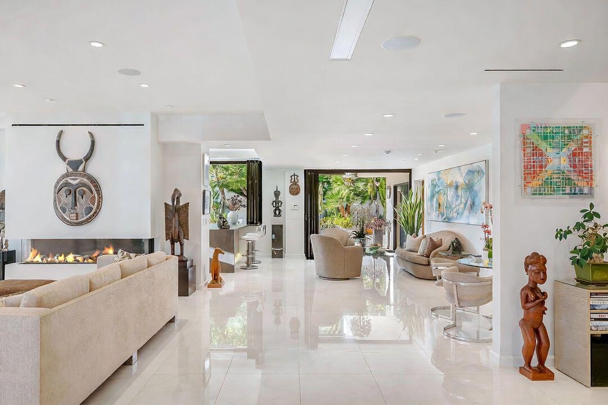 This expansive, open-concept living area features a bright, minimalist aesthetic with polished light-colored flooring and white walls. A prominent fireplace with a linear firebox serves as a focal point, adorned with a large circular tribal-style wall hanging. The space flows seamlessly into a dining or lounge area that opens to a lush outdoor patio, creating a sophisticated and airy atmosphere suitable for high-end entertaining.