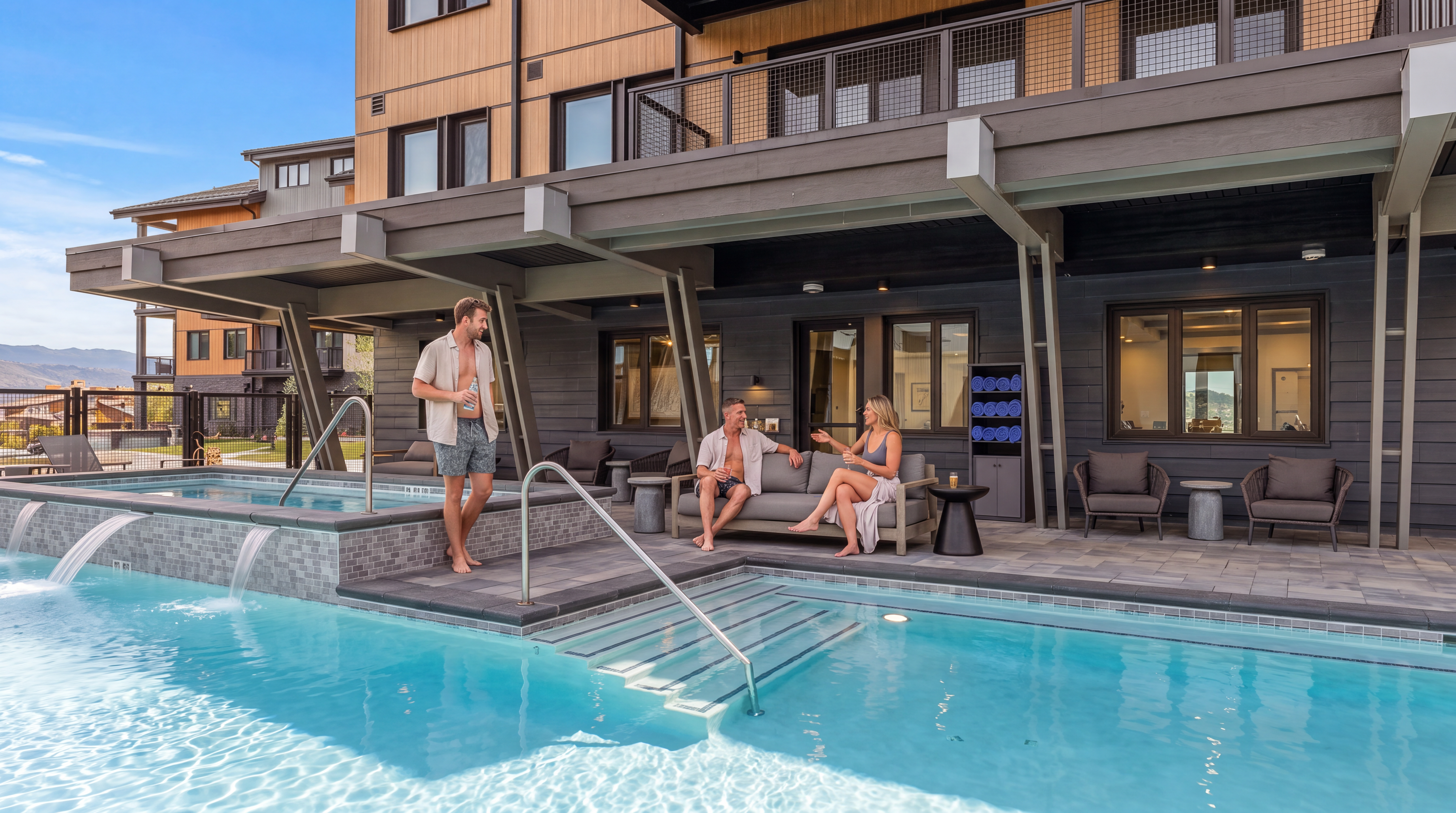 This evening shot captures a luxurious pool and spa area set against a modern multi-story building with dark, textured siding and wood-toned accents. The scene features a glowing, illuminated pool with a cascading water feature from the raised spa, surrounded by comfortable outdoor lounge seating under a covered patio. The perspective is low and centered, emphasizing the inviting, resort-style atmosphere of the community amenity.