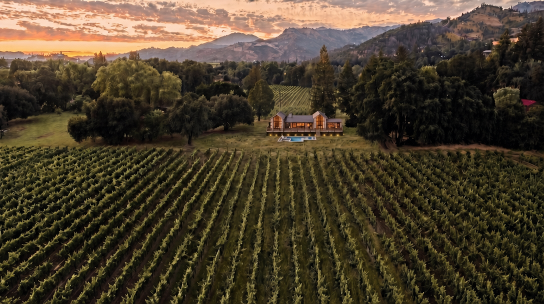 This breathtaking aerial view showcases an elegant house nestled amidst a vast, lush vineyard at sunset. The property features a prominent swimming pool facing the vineyards and rolling mountain ranges in the distance, exuding a luxurious and tranquil atmosphere. The architectural style blends rustic charm with modern refinement, making it an idyllic retreat.