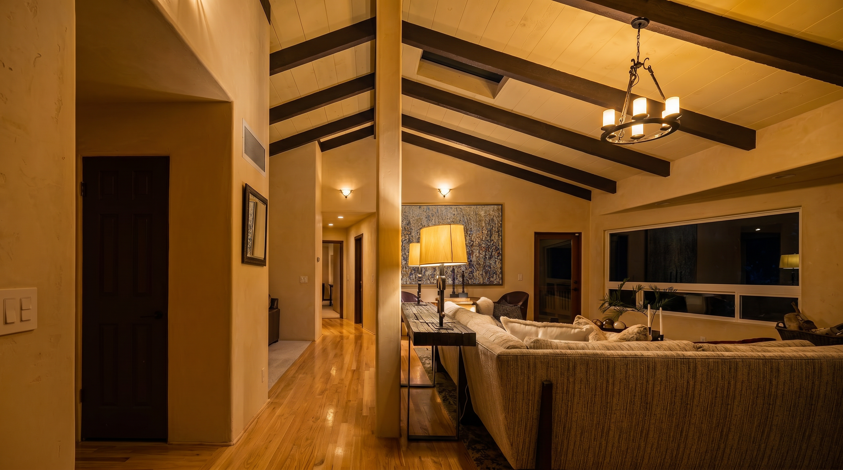 This spacious living room features a dramatic vaulted ceiling with exposed dark wood beams and a central skylight, creating an airy and open atmosphere. A large, light-colored sectional sofa sits on a patterned area rug, complemented by a long, narrow console table and a rustic chandelier. The room flows seamlessly into a hallway with light wood flooring, while a large window provides a view of the lush greenery outside.