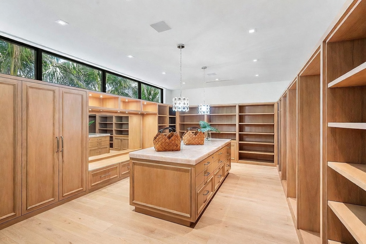 This spacious, high-end walk-in closet features extensive light wood cabinetry and shelving throughout, providing ample storage and organization space. A central island with a stone countertop and multiple drawers sits in the middle of the room, complemented by two decorative pendant lights hanging above. The room is brightened by a long, horizontal window that spans the upper wall, offering a view of lush greenery outside.