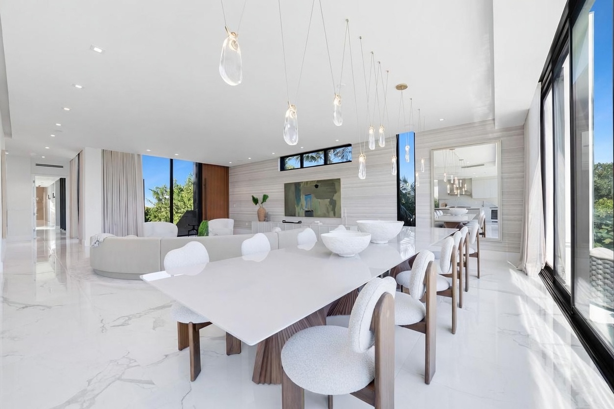 This expansive, open-concept dining and living area features polished light-toned stone flooring and a minimalist aesthetic. A long, sleek dining table is surrounded by curved, textured chairs, while a large, curved sofa defines the adjacent living space. The room is illuminated by a series of unique, organic-shaped pendant lights and benefits from abundant natural light streaming through large windows that overlook lush greenery.