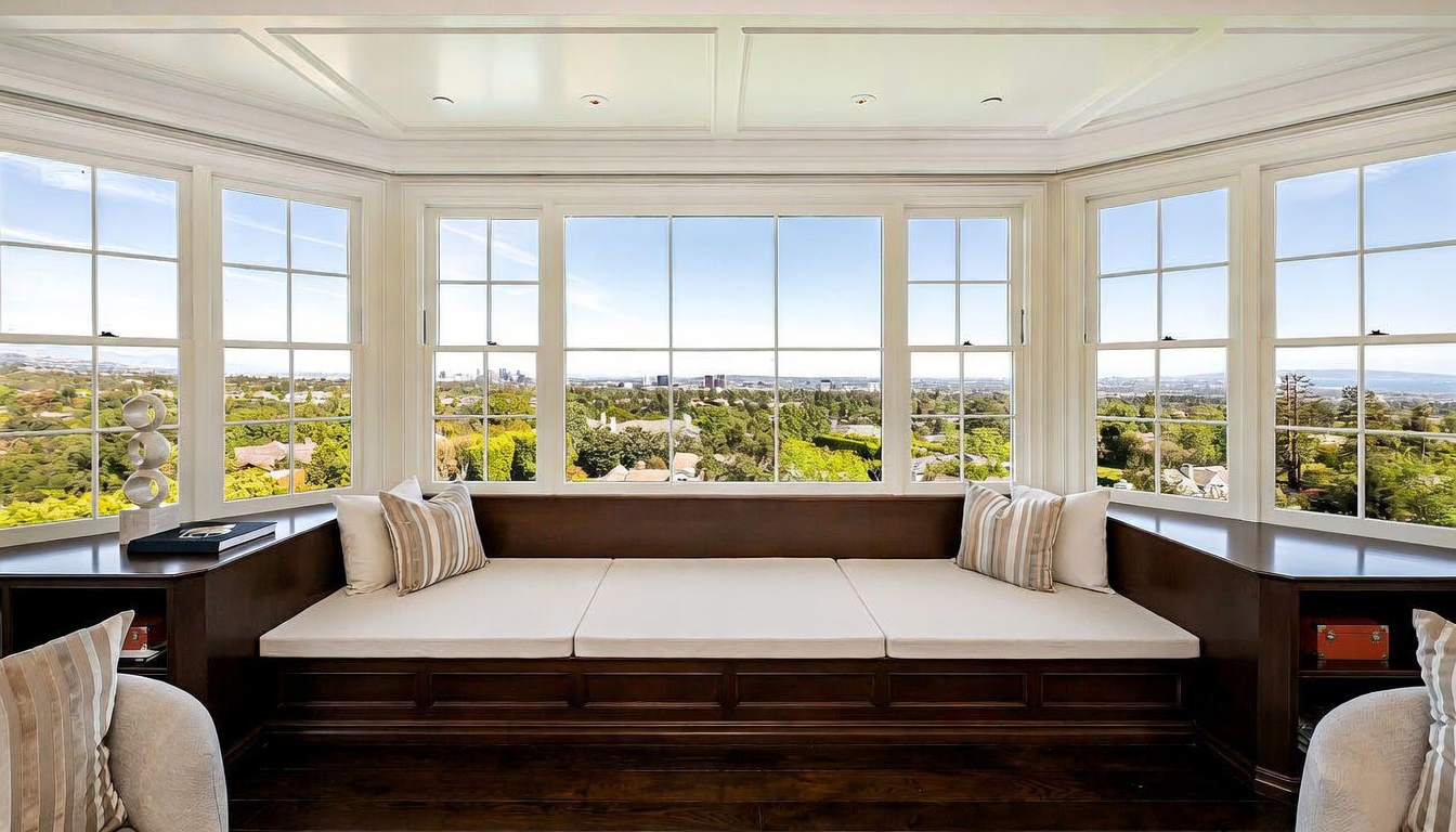 This interior space features a built-in window seat spanning a large bay window, offering expansive panoramic views of the surrounding landscape and city skyline. The dark wood cabinetry of the window seat contrasts with the light-colored cushions and pillows, creating a cozy and inviting reading nook. The room is framed by a coffered ceiling and symmetrical side cabinets, emphasizing a refined and classic architectural style.