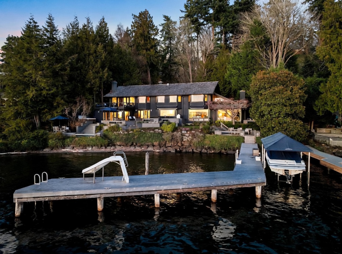 This stunning waterfront property features a dark-toned, multi-level home nestled against a lush, forested hillside. The exterior showcases a spacious stone patio, a private wooden dock with a boat lift, and a slide, all overlooking a serene lake. The perspective is captured from the water, highlighting the seamless integration of luxury living with the natural landscape.