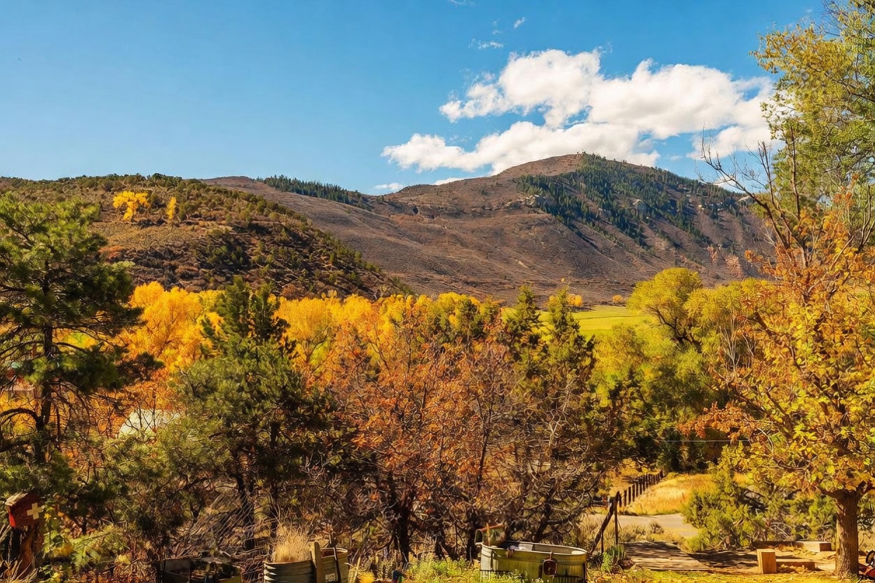 This scenic outdoor view captures a vibrant autumn landscape with golden-hued trees set against a backdrop of rolling mountains under a clear blue sky. The foreground features a rustic, natural setting with a metal trough and a wooden fence, suggesting a peaceful, rural property. The perspective is an eye-level shot looking out over the expansive valley, emphasizing the tranquil and picturesque nature of the location.