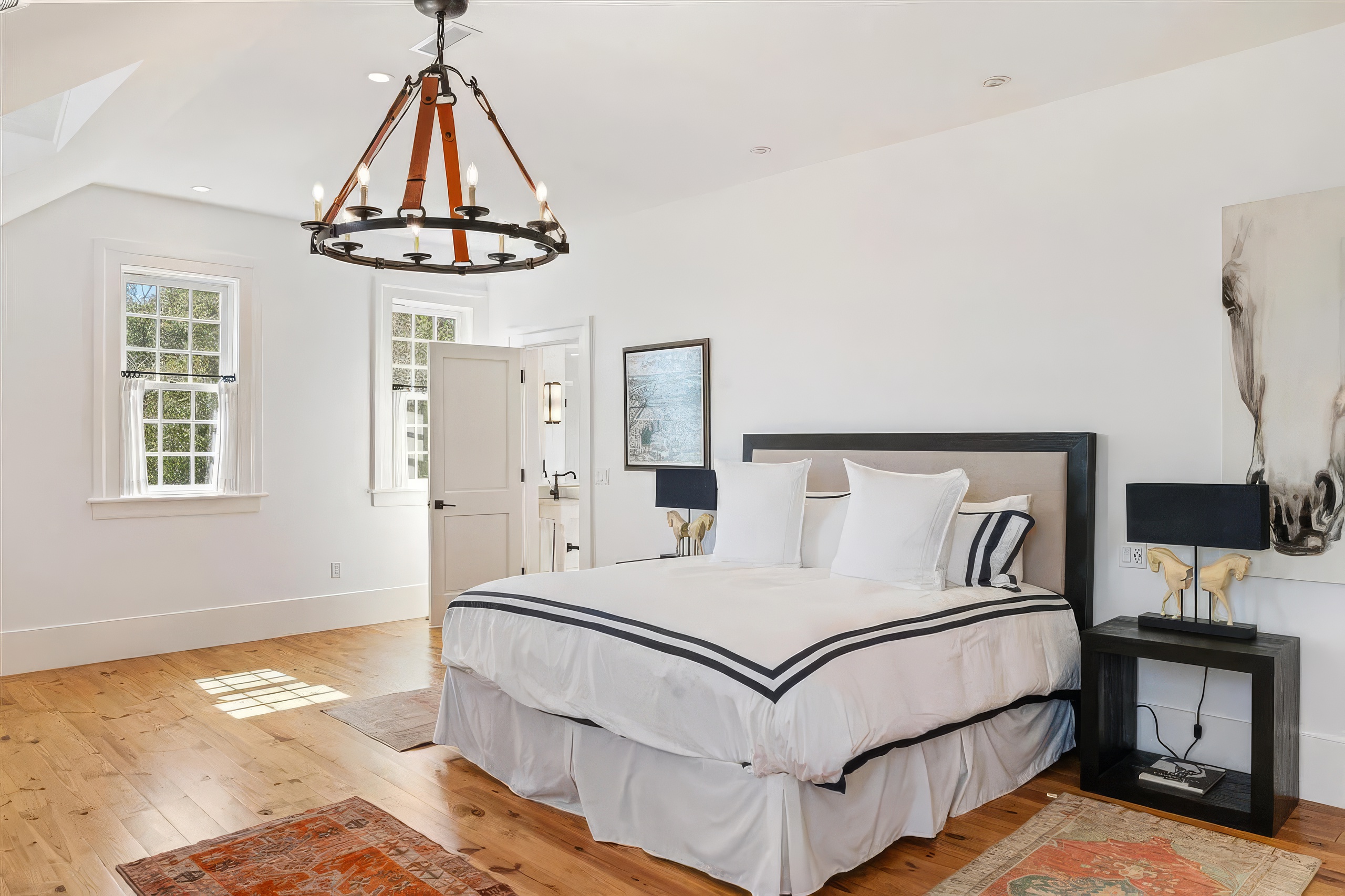 This bright and airy primary bedroom features light wood flooring, white walls, and a large central bed with a dark-framed headboard. A distinctive circular chandelier with leather straps hangs from the ceiling, while two black bedside tables with matching lamps provide a modern contrast. The room is accented by a large horse-themed artwork and two patterned rugs, creating a sophisticated and welcoming atmosphere.