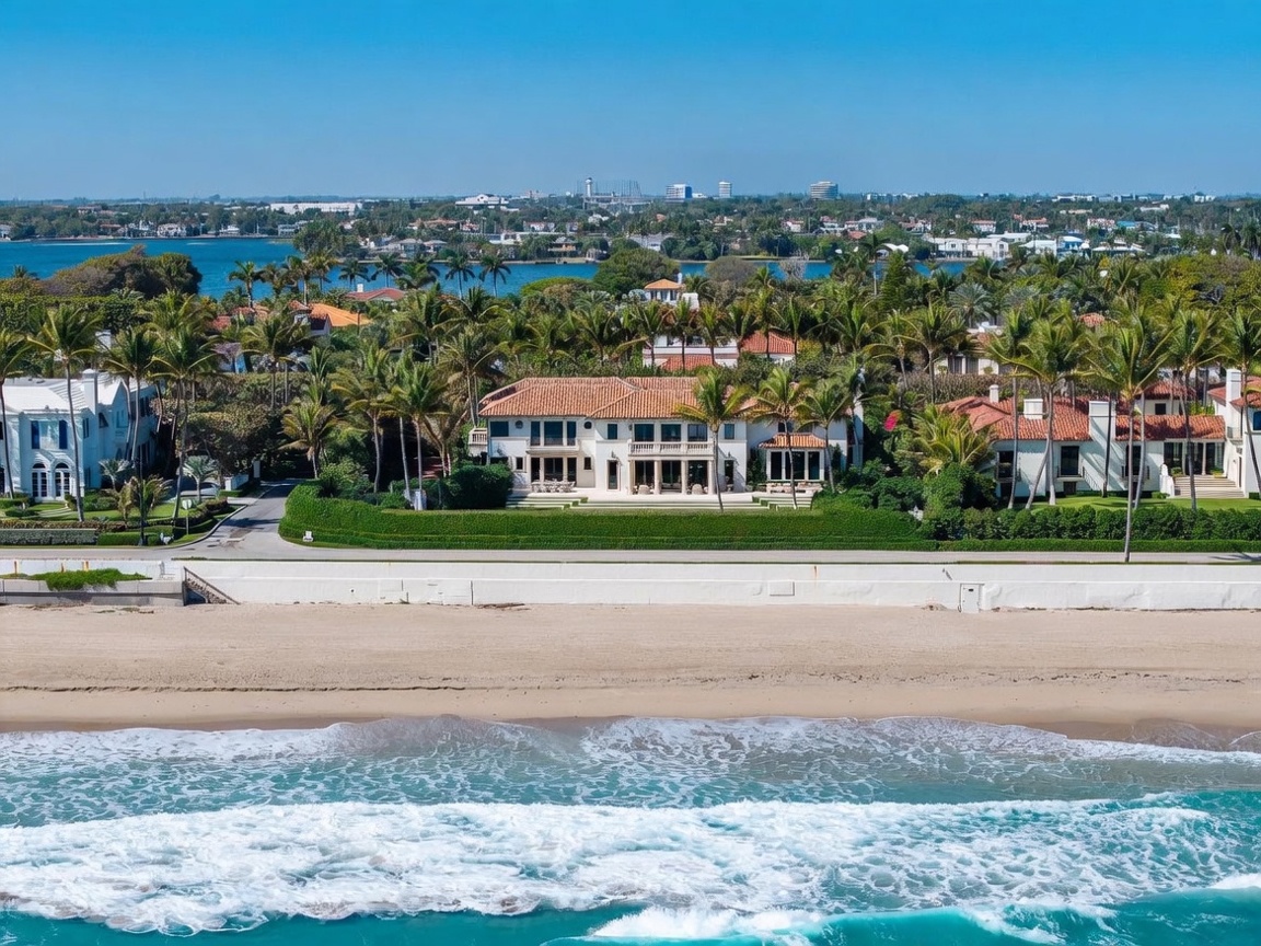 This high-angle aerial shot captures a luxurious beachfront estate featuring a Mediterranean-style residence with a terracotta tile roof and a symmetrical facade. The property is nestled between a pristine sandy beach and a lush, palm-filled landscape, with a scenic waterway visible in the background. The perspective emphasizes the home's prime coastal location, showcasing its expansive lawn, manicured hedges, and direct access to the ocean.