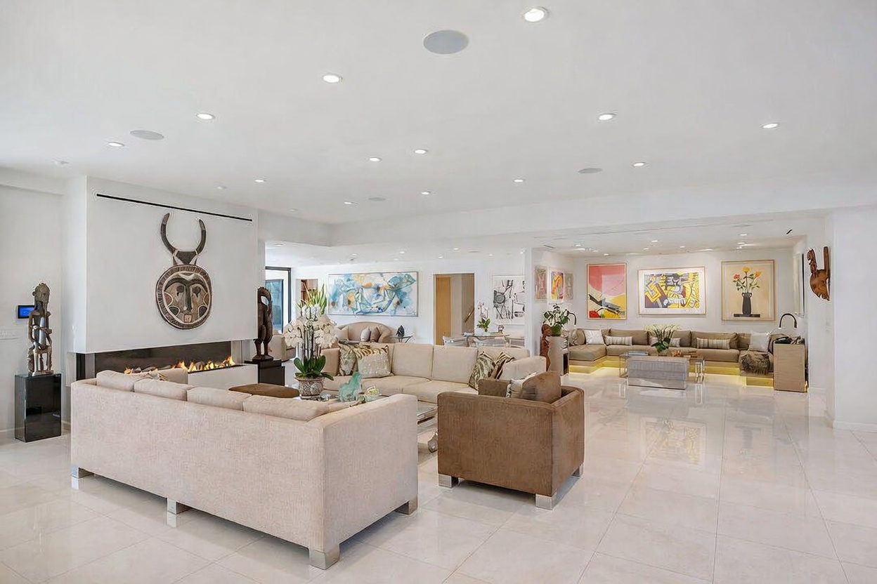 This expansive, open-concept living area features polished light-colored stone flooring and a bright, neutral color palette. The space is anchored by a contemporary fireplace with a prominent, tall, dark-toned surround adorned with decorative wall art, while multiple seating areas are arranged with plush sofas and armchairs. The room is illuminated by numerous recessed ceiling lights and accented with various framed artworks, creating a sophisticated and airy atmosphere.