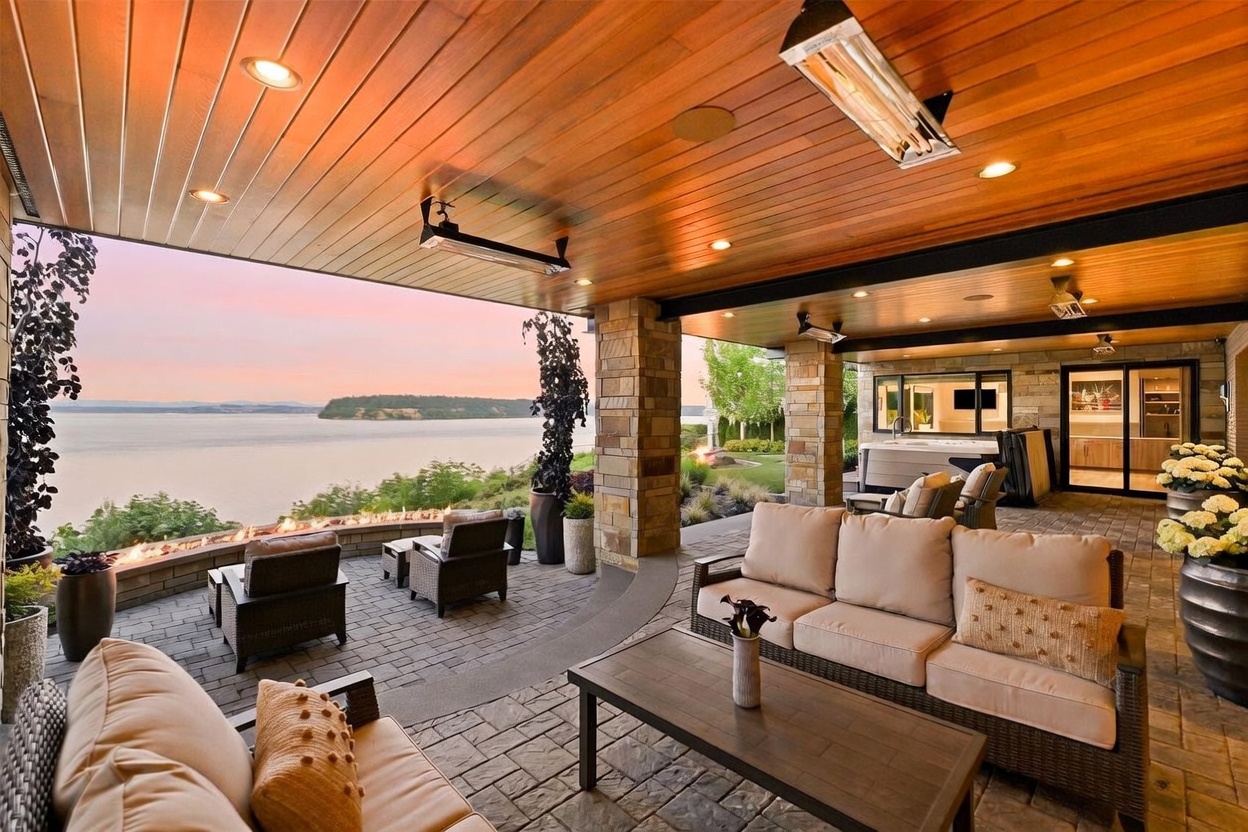 This expansive covered patio offers a luxurious outdoor living space with a panoramic view of the water at sunset. The area features comfortable lounge seating, a linear fire feature built into a stone wall, and a hot tub tucked into the corner, all sheltered by a warm wood-paneled ceiling with integrated heaters. The stone-paved floor and natural stone pillars create a sophisticated, high-end atmosphere perfect for evening relaxation.
