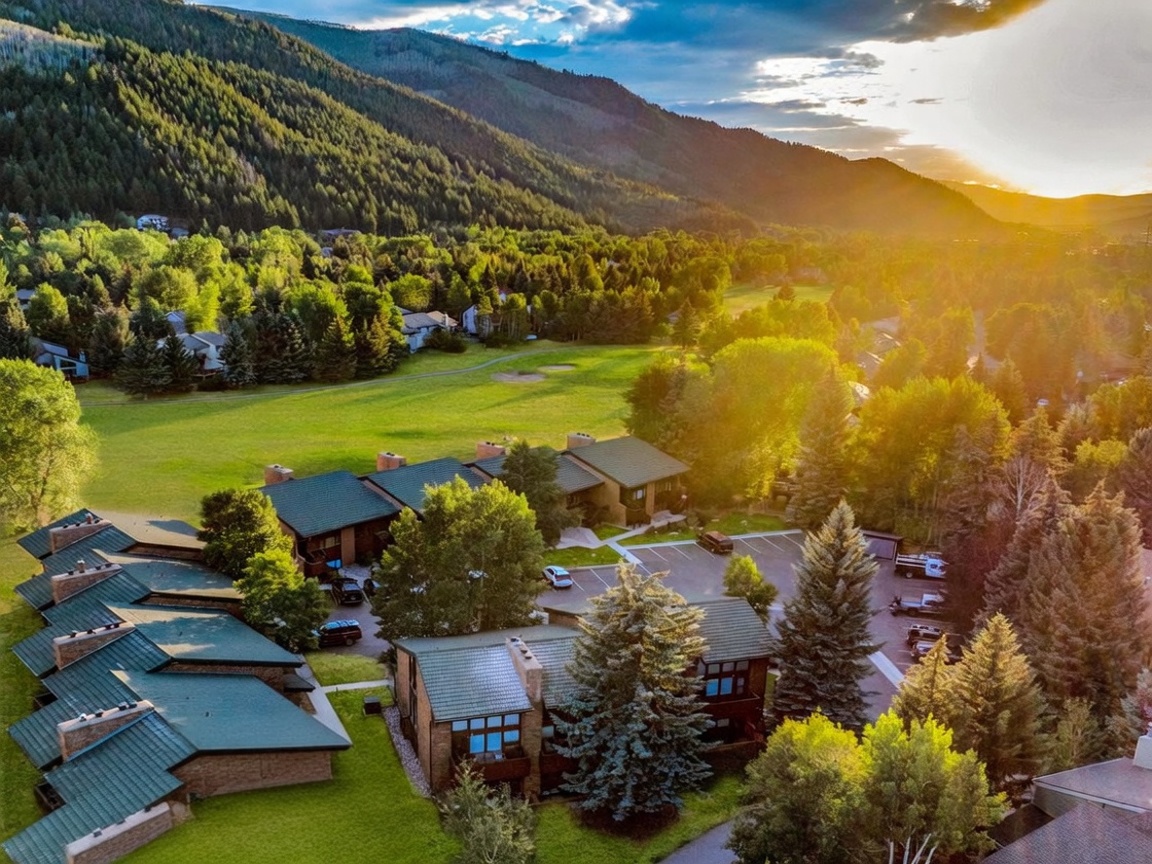 This aerial view showcases a peaceful residential complex set against a stunning mountain backdrop during golden hour. The property features townhome-style residences with green shingled roofs, a well-maintained lawn, and convenient surface parking. The expansive surrounding green space and mature trees create a scenic and tranquil atmosphere ideal for a mountain retreat.
