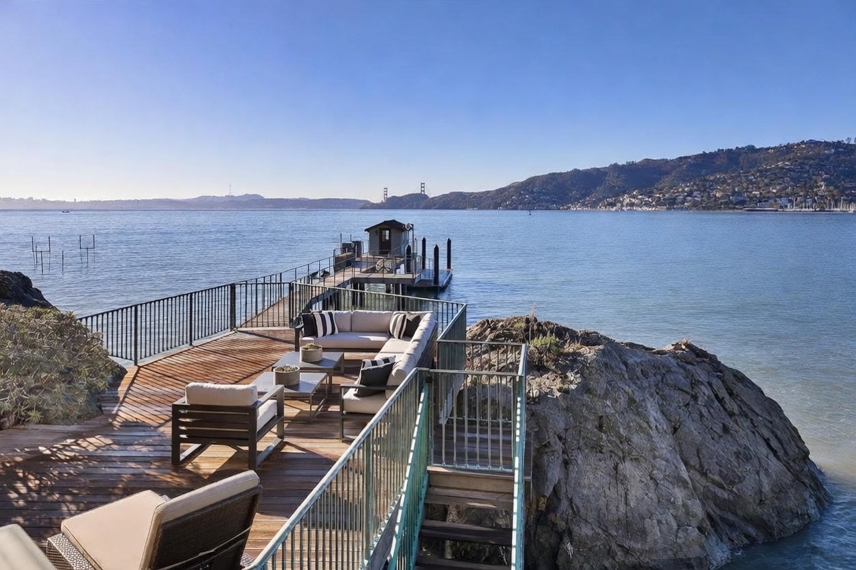 This stunning waterfront deck offers a luxurious outdoor living space perched directly above the bay, featuring comfortable modern lounge seating and a wooden plank surface. The deck extends into a private pier leading to a small boat house, with panoramic views of the distant coastline and the Golden Gate Bridge. The perspective captures the seamless integration of the home's architecture with the natural rocky shoreline and the expansive water.