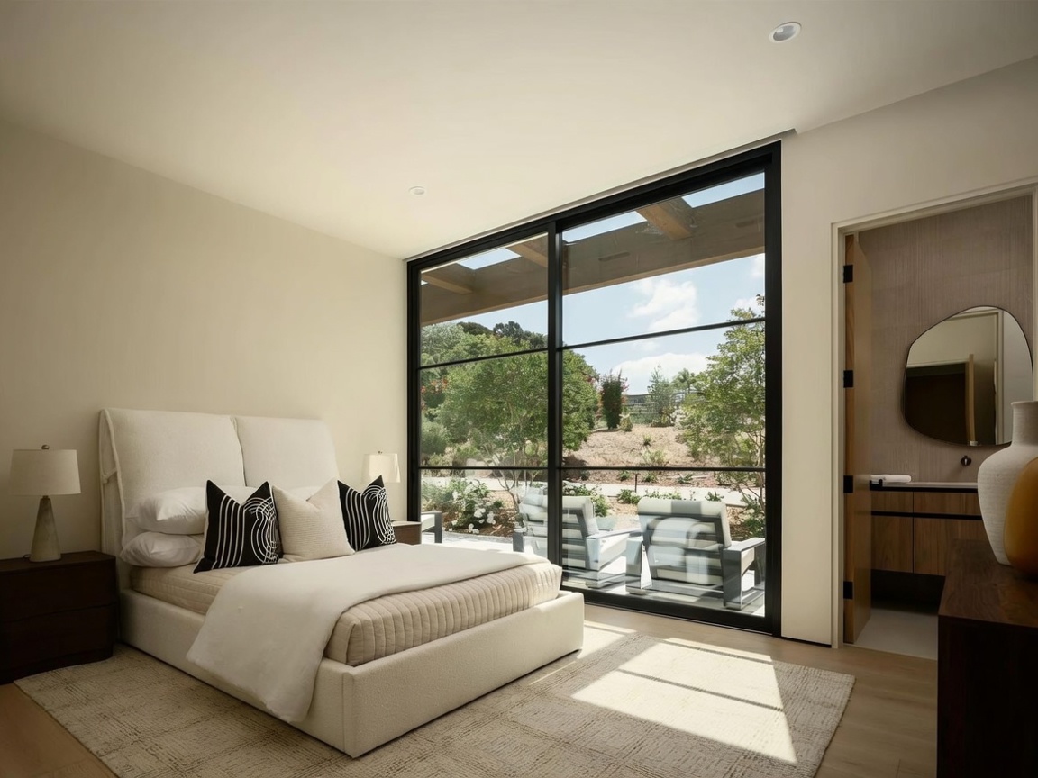 This bright and airy primary bedroom features a neutral color palette and a large, floor-to-ceiling window wall that provides a seamless connection to the outdoor patio. The room is furnished with a plush upholstered bed, dark wood nightstands, and a textured area rug, while an adjacent open doorway leads into a modern bathroom with a unique organic-shaped mirror. The overall aesthetic is minimalist and sophisticated, emphasizing natural light and a tranquil indoor-outdoor flow.