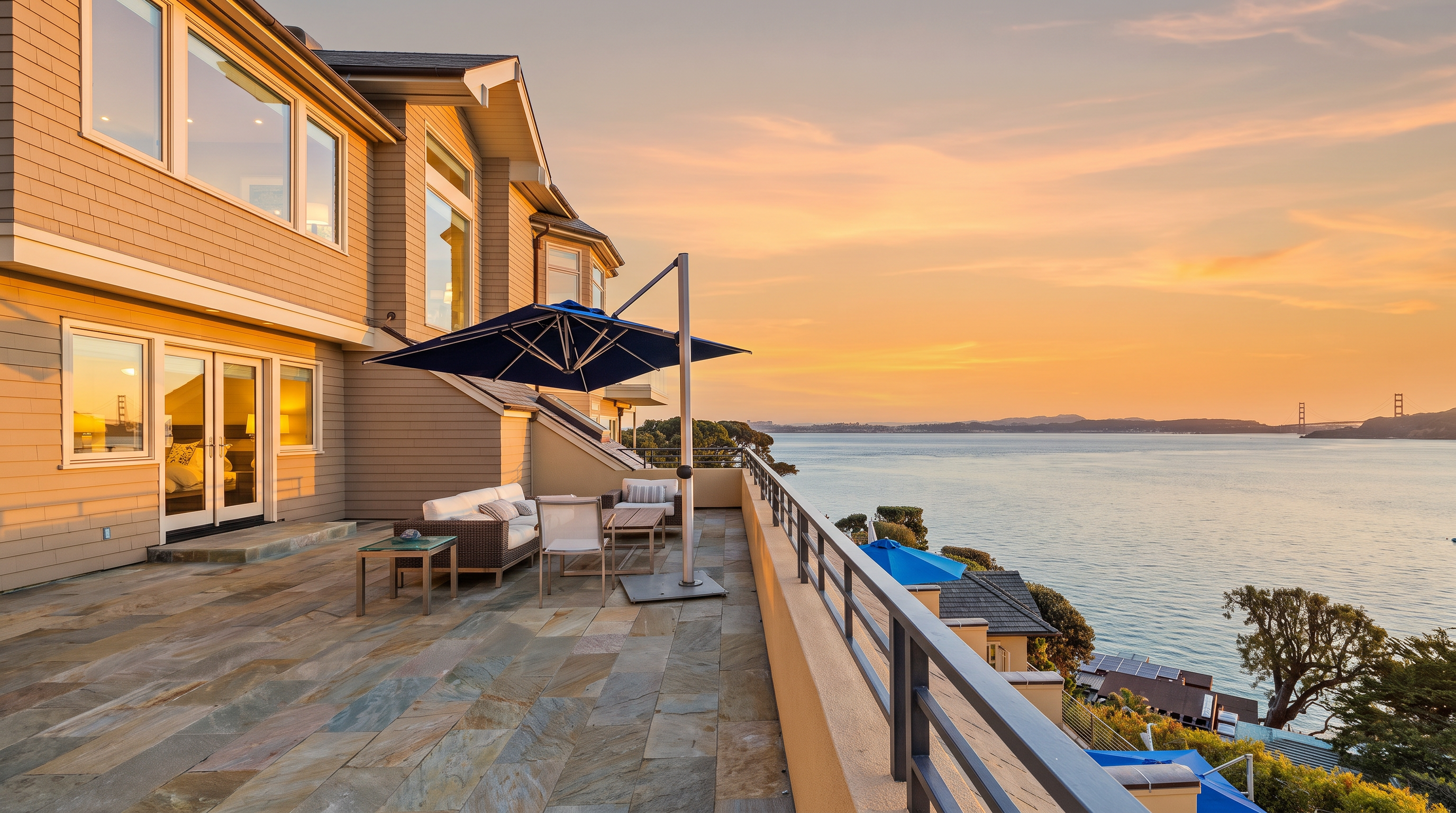 This expansive, stone-paved balcony offers a breathtaking panoramic view of the bay, featuring a clear sightline to the Golden Gate Bridge in the distance. The space is furnished with comfortable outdoor seating and a large cantilevered umbrella, creating an ideal setting for relaxation or entertaining. The scene is captured from a slightly elevated perspective, emphasizing the seamless transition between the home's exterior and the vast, serene waterfront.