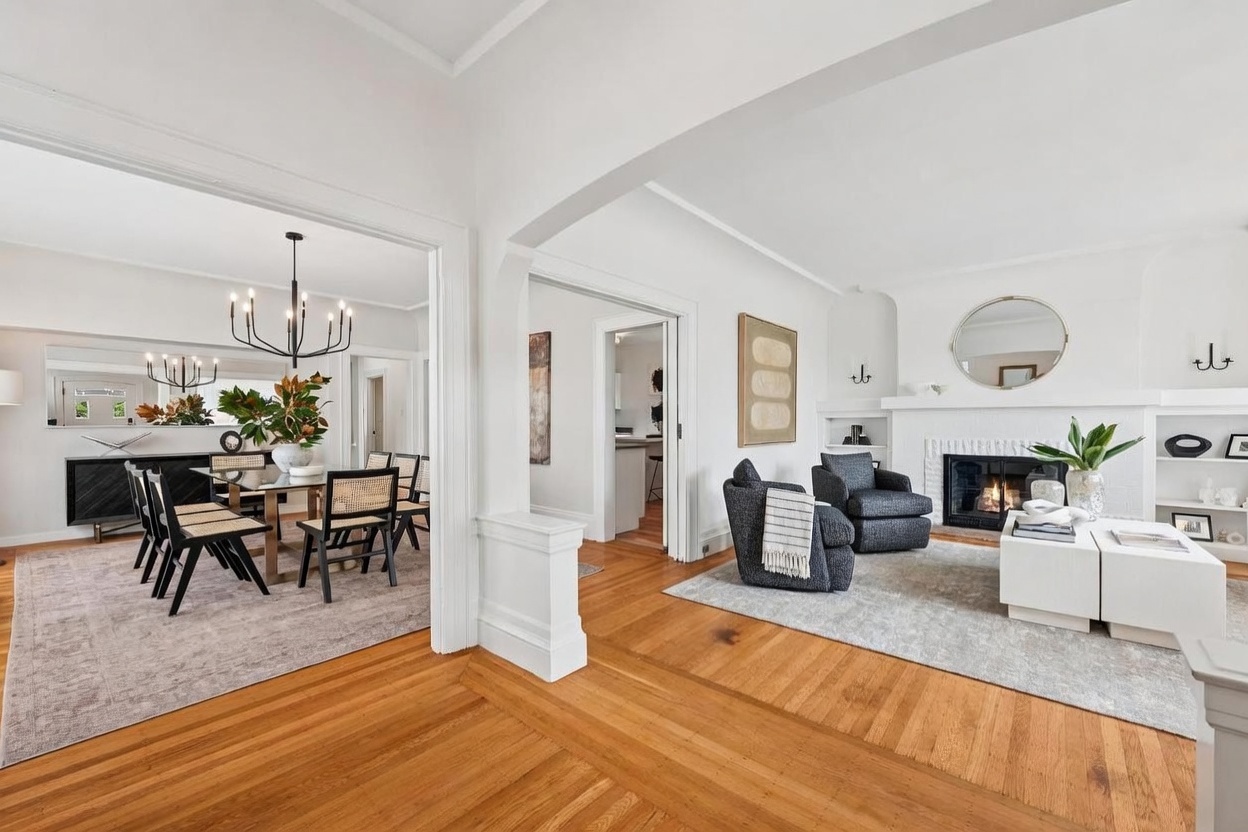 This bright and airy open-concept living space features warm hardwood flooring and crisp white walls, creating a seamless flow between the living and dining areas. The living room is anchored by a white brick fireplace surround with built-in shelving, while the dining room is highlighted by a modern black chandelier and a large mirror. The perspective is captured from a central vantage point, showcasing the elegant transition between rooms and the sophisticated, neutral decor.