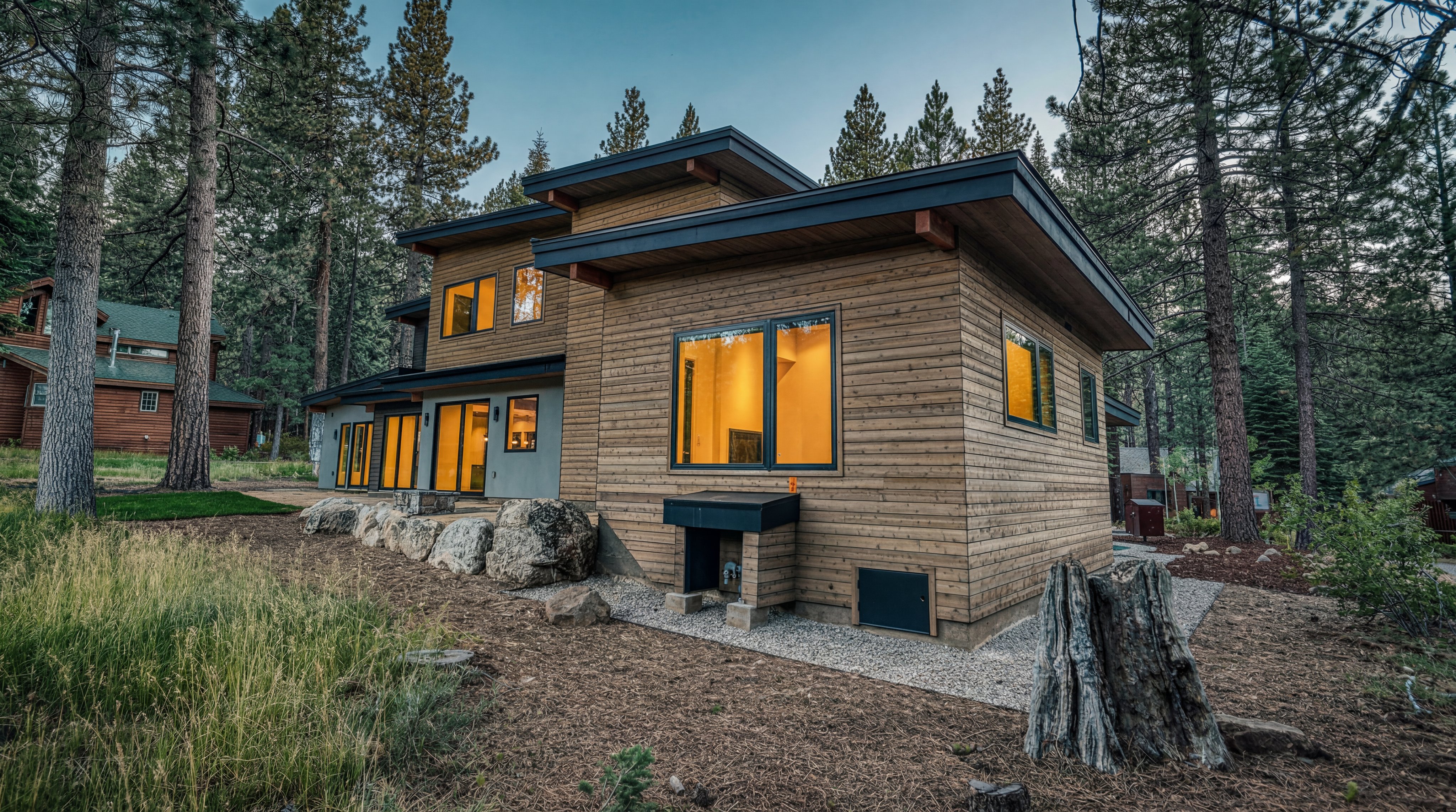 This modern mountain home features horizontal wood siding and a flat, overhanging roofline, set against a backdrop of tall pine trees. The exterior includes large windows that glow with warm interior light, contrasting with the natural, rugged landscape of boulders and pine needles. The perspective captures the side profile of the structure, emphasizing its contemporary architectural design and integration into the forested environment.