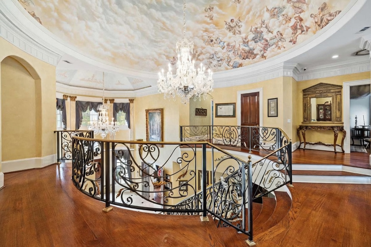 This grand upper-level landing features a dramatic curved wrought-iron railing overlooking the foyer below. The space is defined by a stunning hand-painted mural on the domed ceiling, complemented by an ornate crystal chandelier and warm wood flooring. The architectural style is opulent and classical, creating a sophisticated and airy atmosphere.
