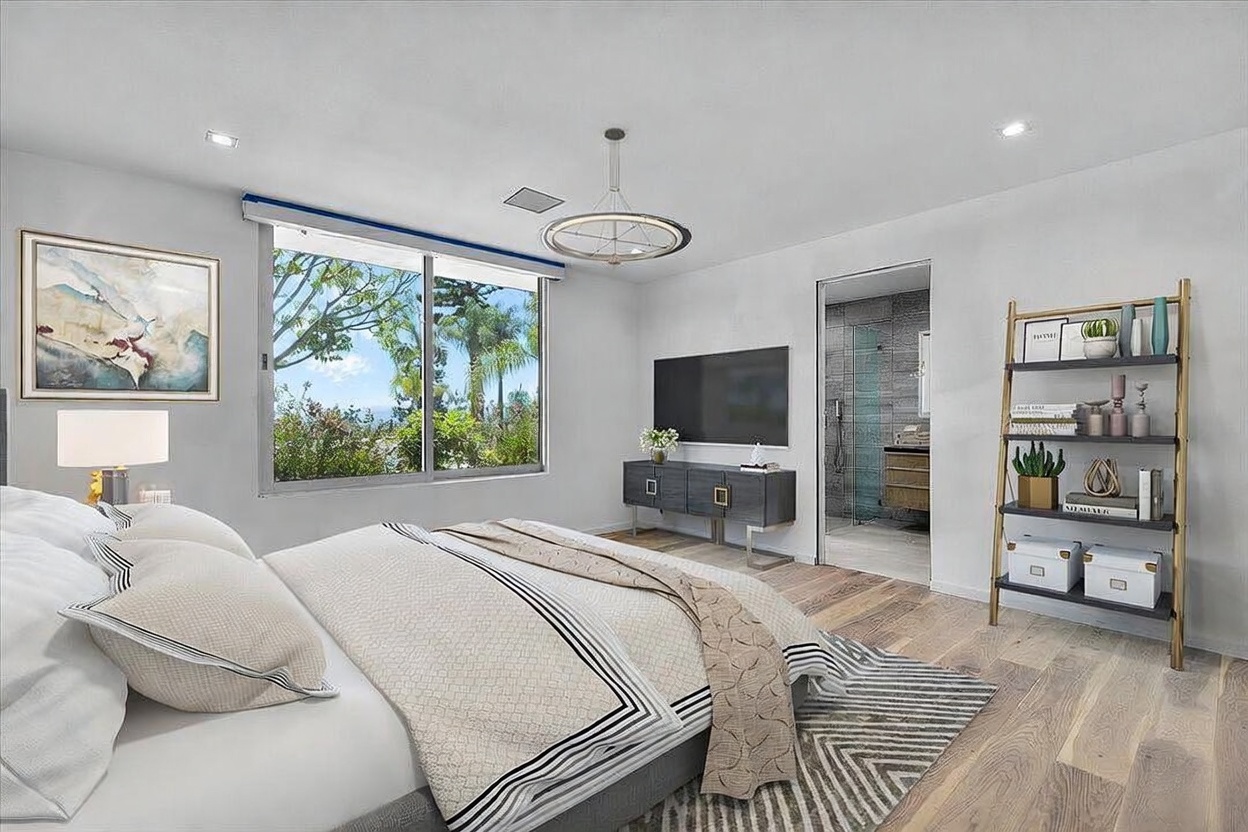This bright and airy primary bedroom features light wood flooring and a neutral color palette, creating a serene atmosphere. A large window provides ample natural light and a view of the lush greenery outside, while a modern chandelier adds a touch of elegance. The room is furnished with a comfortable bed, a sleek media console beneath a wall-mounted television, and a decorative shelving unit, with an open doorway leading into an adjacent bathroom.